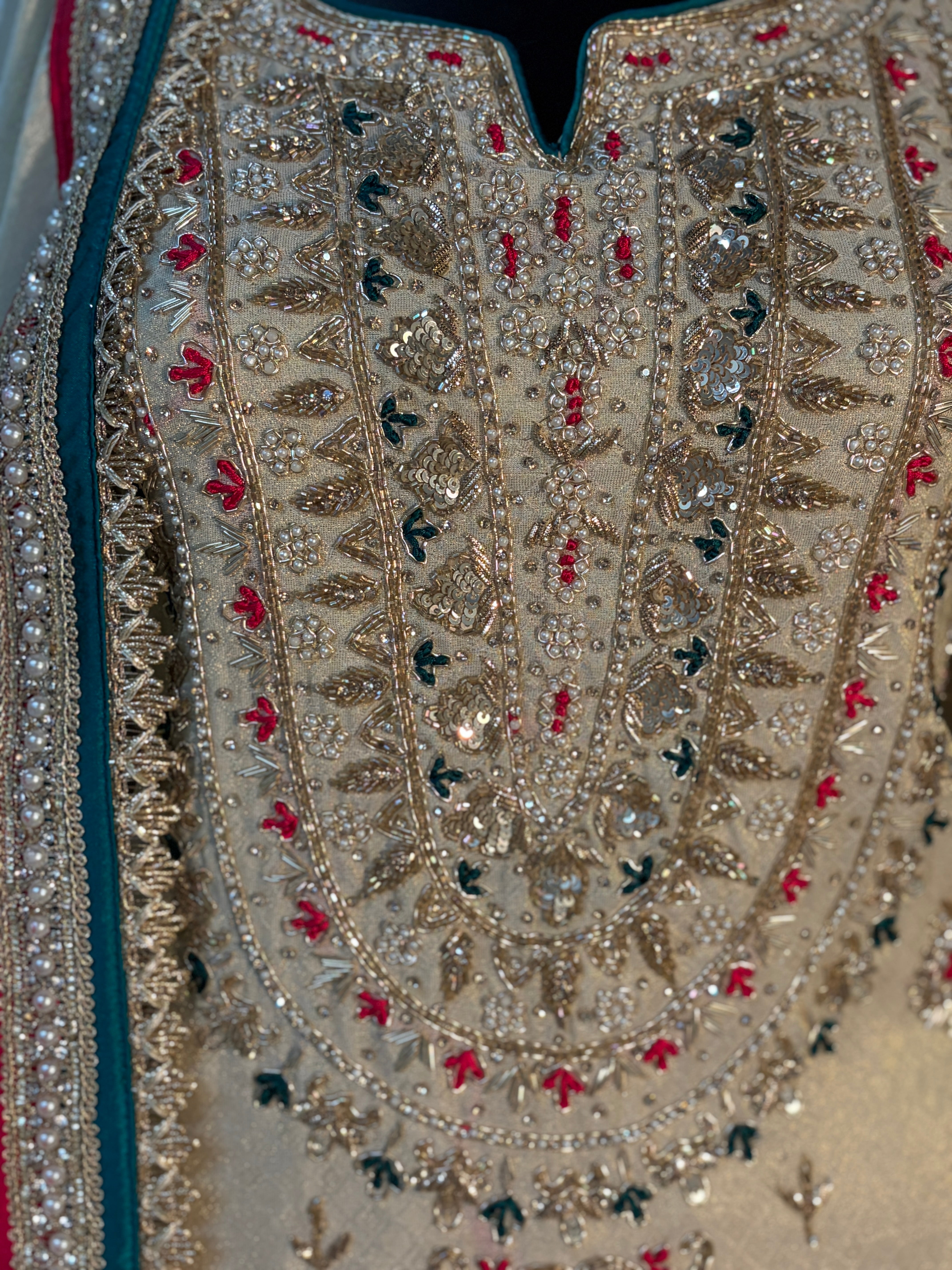 Close-up of an ornate beige embroidered garment with intricate metallic gold beadwork, silver mirror work, and small red and dark green embroidered accents.