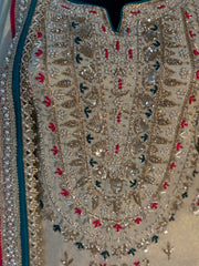 Close-up of an ornate beige embroidered garment with intricate metallic gold beadwork, silver mirror work, and small red and dark green embroidered accents.